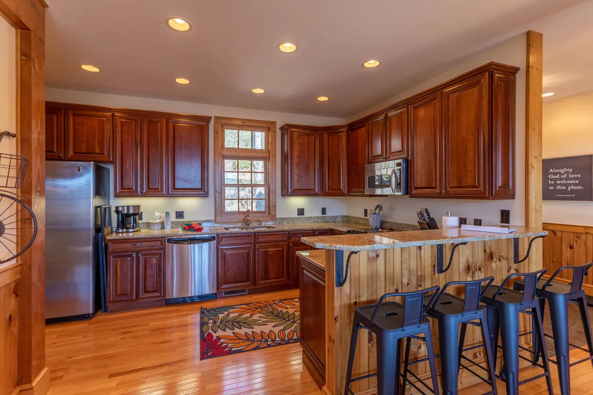 A cozy and inviting kitchen space with a large island, wooden cabinetry, stainless steel appliances, and a colorful area rug. The open format makes it easy for multiple people to cook, gather, and interact, supporting the idea of group cooking and shared experiences. The design is senior-friendly and accessible, with plenty of space for movement.