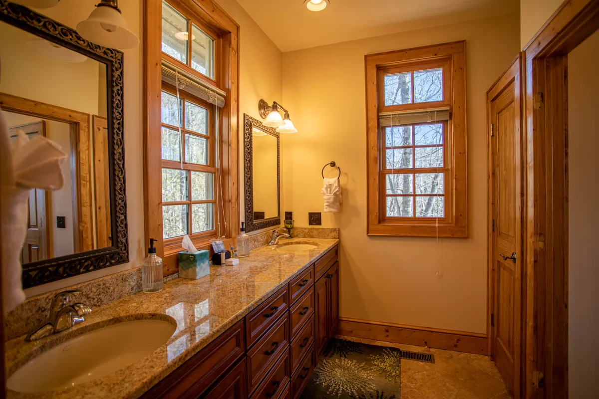 A bright, elegant bathroom with double sinks set in a granite countertop, large mirrors reflecting natural light, and windows that create a tranquil, refreshing ambiance. The space is both stylish and functional, ideal for shared or private routines.