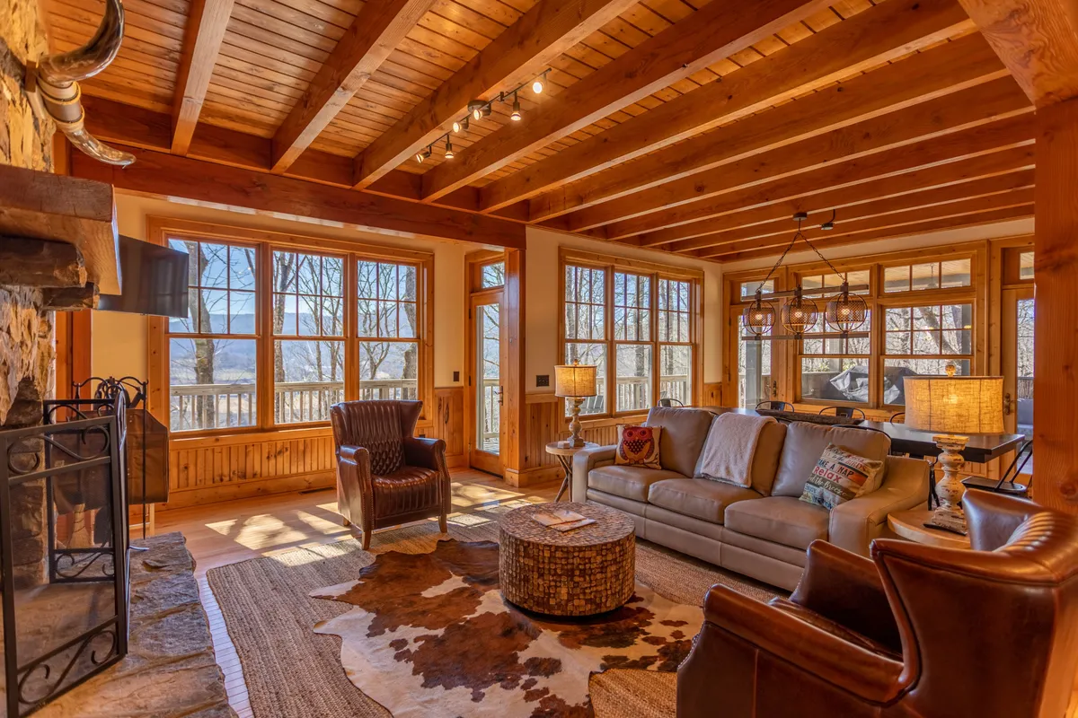 A spacious living area with large windows letting in abundant natural light, comfortable seating, a stone fireplace, rustic wooden beams, and a cowhide rug. The windows offer glimpses of the surrounding mountain landscape, connecting the interior to the breathtaking scenery outside.
