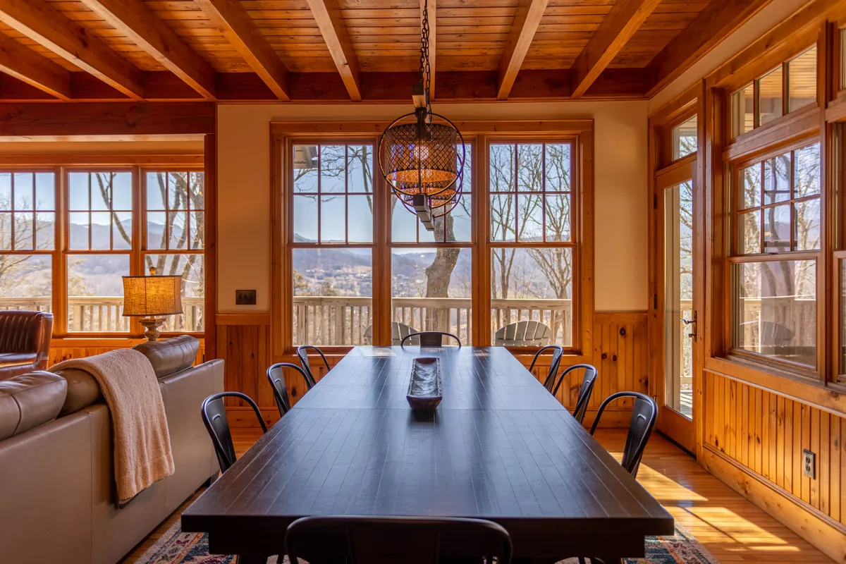 A long dining table set beneath a chandelier, flanked by large windows that frame the mountains beyond. The cozy setting is ready for family dinners or celebrations, with ample space for a group to gather.