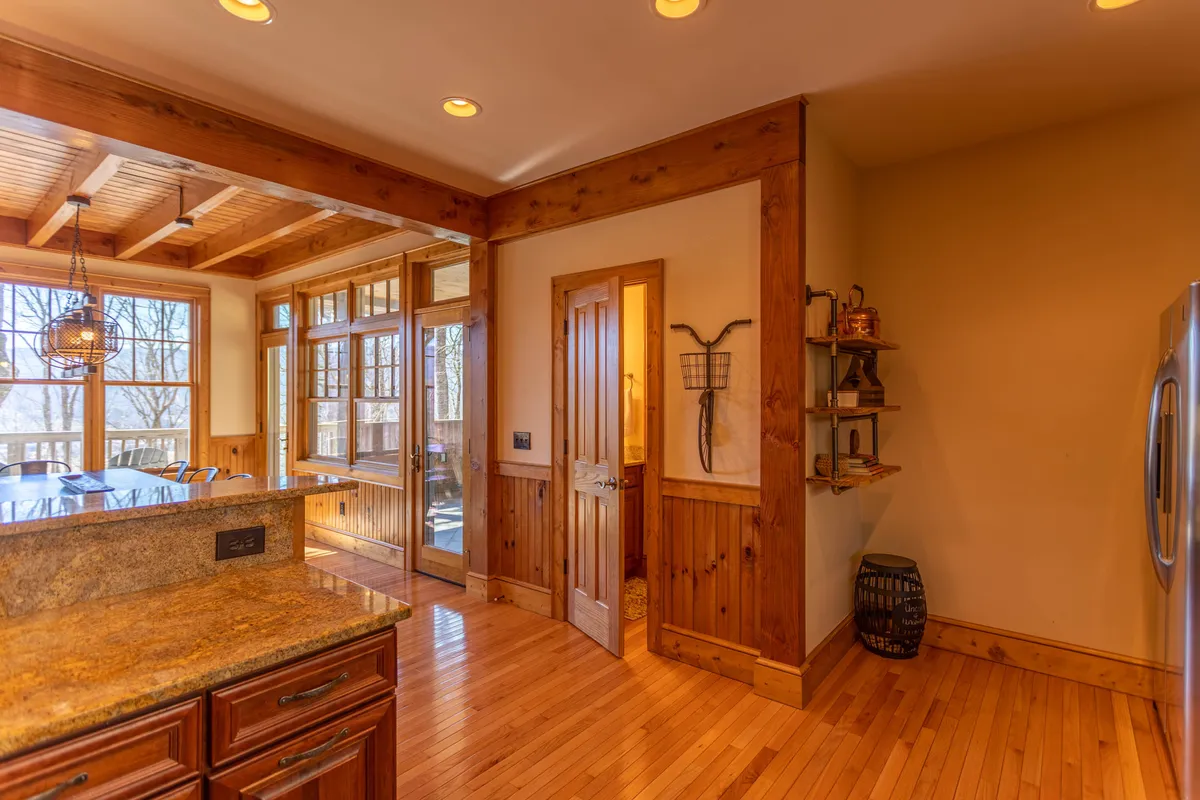 An open-plan kitchen and dining area with a prominent kitchen island and wooden cabinetry, surrounded by large windows letting in natural light. The adjacent dining table is set for meals, blending cooking and gathering spaces with views outside.