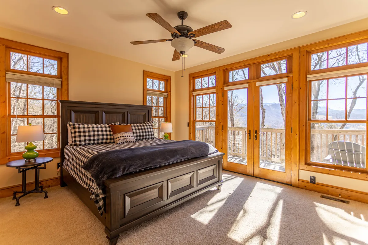 A tranquil bedroom with a comfortable bed positioned by large windows that let in ample natural light. The space features a ceiling fan and a nightstand with a lamp, creating a serene atmosphere perfect for relaxation and privacy.