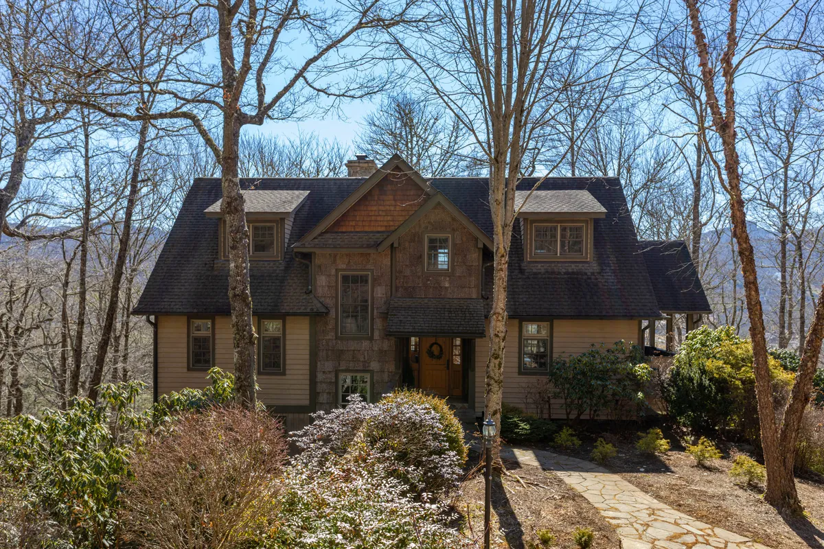 The image showcases the inviting facade of a craftsman-style single-family house nestled among mature trees, with a stone pathway leading to a spacious porch. The porch offers sweeping sunset views and is decorated seasonally, setting a warm, tranquil tone. The lush greenery and gentle light evoke a peaceful, secluded retreat, while hints of the porch's comfort promise relaxing mornings with coffee amid nature.