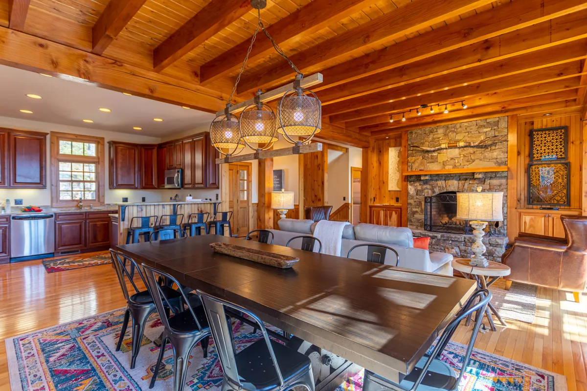 This image showcases a large wooden dining table surrounded by stylish light fixtures, a stone fireplace, and an open kitchen area. The space is clearly designed for gatherings, with ample seating and a welcoming, open layout that encourages conversation and shared meals. The visible kitchen hints at the dual kitchen setup referenced in the section, and the overall ambiance is warm and inviting.