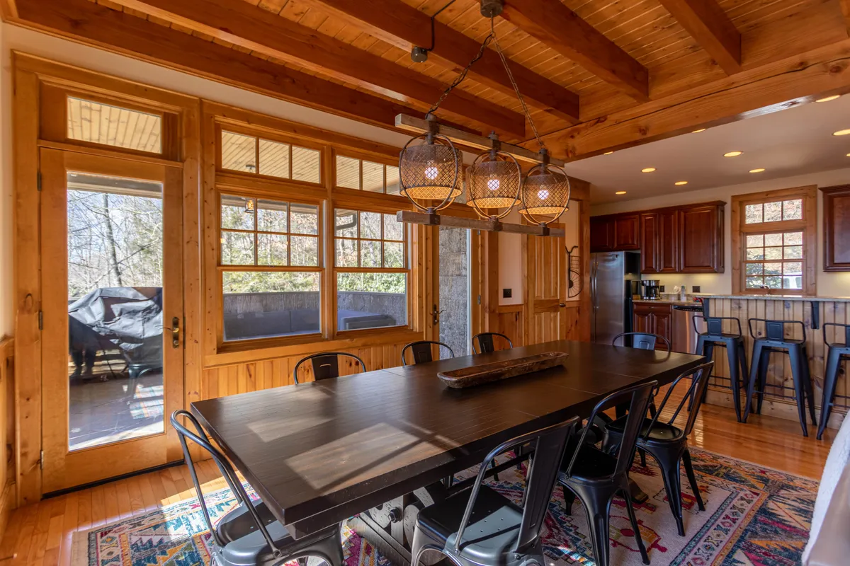 Features a large dining table beneath hanging light fixtures, wooden beam ceiling, and windows letting in natural light. The setting radiates comfort and hospitality, making it an ideal spot for festive gatherings or quiet moments with family across generations. The bright and airy feel, with accessible main-level living, supports the notion of accommodating multi-generational needs.