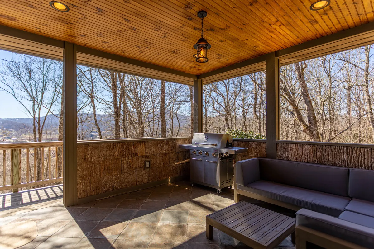 An exterior view of a spacious outdoor grill and seating area with a wooden ceiling and scenic views of trees. The ambiance is relaxed and communal, ideal for barbecues, morning coffee, or evening get-togethers.