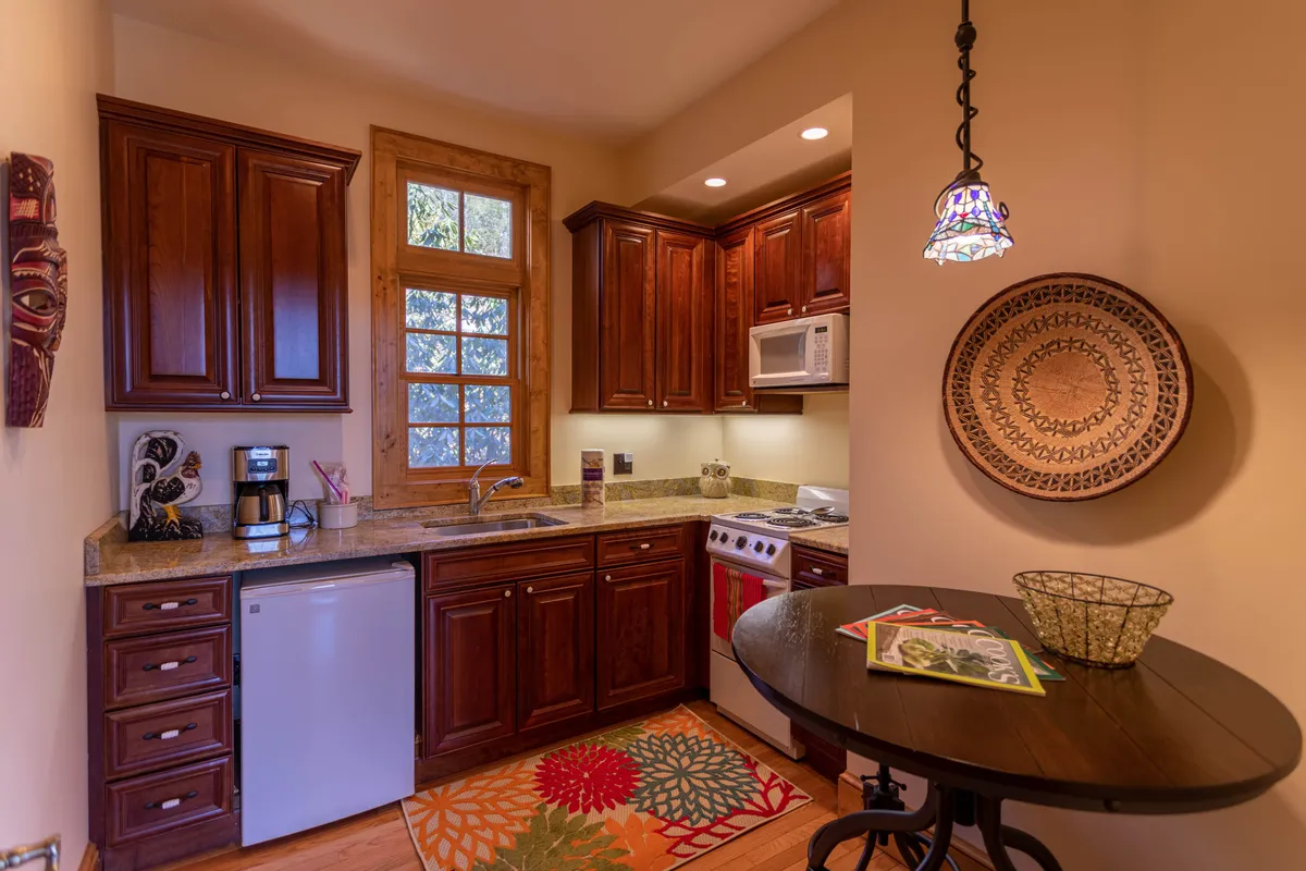 A cozy lower-level kitchen with wooden cabinets, a granite countertop, decorative plate on the wall, colorful rug, and a small table with magazines. The space is inviting and functional, suitable for morning coffee, snacks, or quiet gatherings.