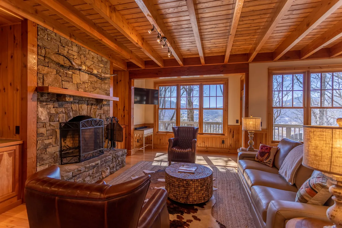 A warm, inviting living room with a stone fireplace, large windows revealing mountain views, comfortable seating, and exposed wooden beams. Sunlight fills the space, and the arrangement invites gatherings and cozy relaxation.